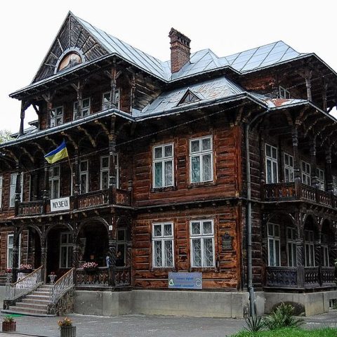 Willa Goplana w Truskawcu, Ukraina, drewniany budynek z elementami stylu zakopiańskiego, z ukraińską flagą i napisem 'Muzeum' nad wejściem.