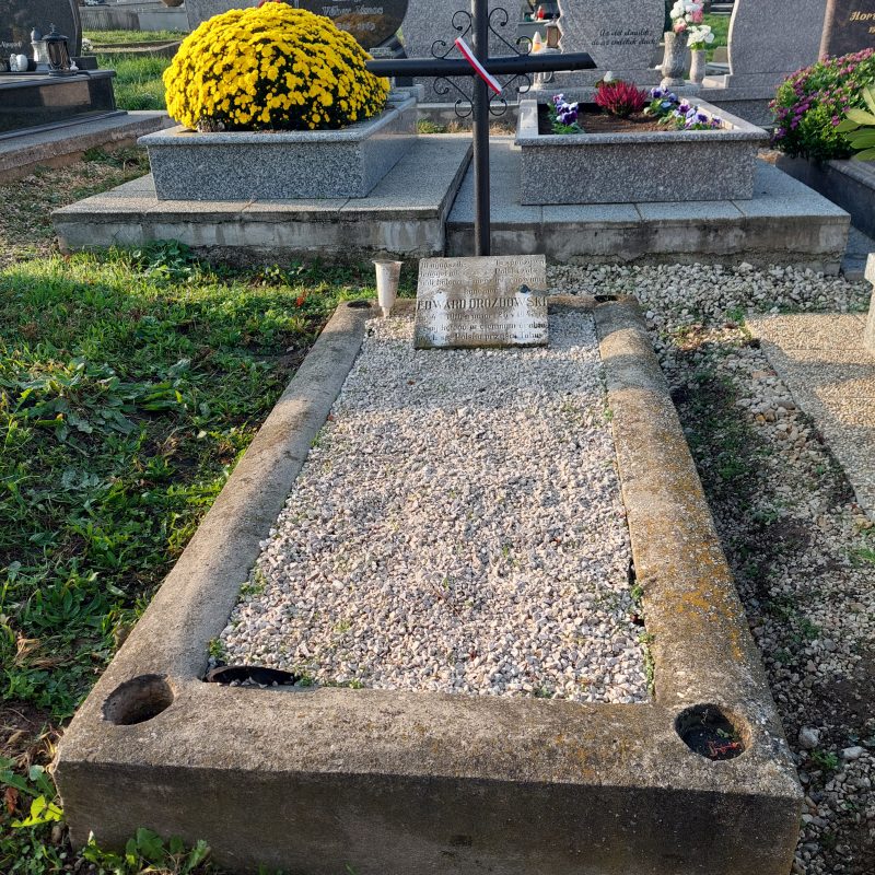 La tombe du soldat polonais Edward Drozdowski, ornée d'une croix métallique et d'une plaque bilingue. La tombe est entourée d'autres pierres tombales et d'un buisson de chrysanthèmes jaunes.