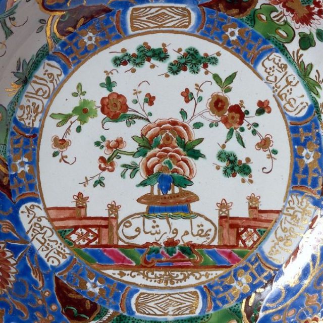 Gros plan sur une assiette de la série « sultan » de la Manufacture royale de faïence du Belvédère, avec un bouquet de fleurs au centre, entouré d'une écriture coufique et de décorations raffinées dans les tons de bleu, de rouge et d'or.