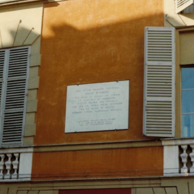 Une plaque commémorative apposée sur un bâtiment orange à Reggio nell’Emilia, en Italie, rendant hommage à la composition de l’hymne national polonais par Józef Wybicki en 1797. La plaque se trouve entre deux fenêtres munies de volets.