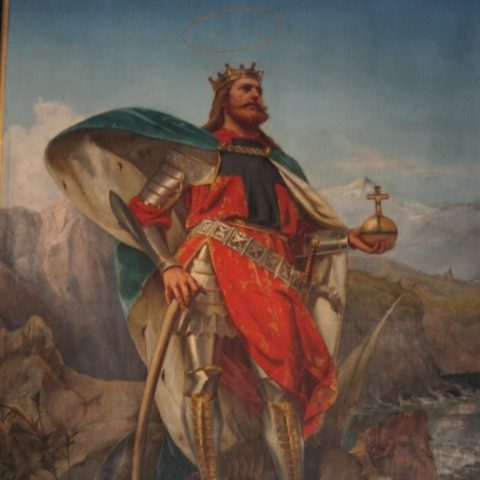 Obraz króla Olafa II w średniowiecznym stroju i zbroi, stojącego heroicznie na smoku na tle skalistego krajobrazu nadmorskiego. Na czarnej ramie znajduje się łacińska inskrypcja.