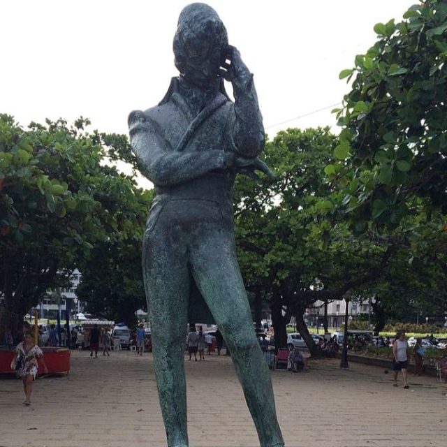 Brązowy pomnik Fryderyka Chopina na niskim postumencie, otoczony drzewami, zlokalizowany na promenadzie przy plaży w Rio de Janeiro. Posąg przedstawia Chopina w zamyślonej pozie.