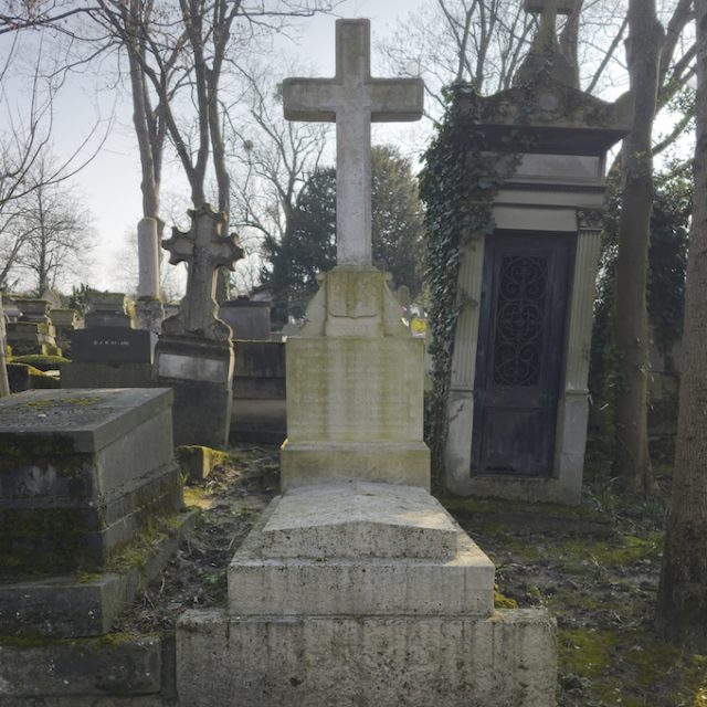 A gravestone with a tall cross on a stepped plinth, with a coat of arms shield. Surrounding graves and trees are visible in the background.