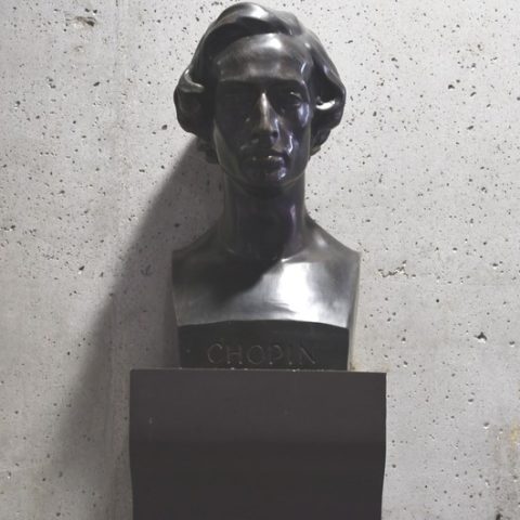 Popiersie Fryderyka Chopina z brązu na betonowej ścianie w Pasażu Chopina w Lublanie. Popiersie ma szczegółowe rysy twarzy i napis 'Chopin' na podstawie.