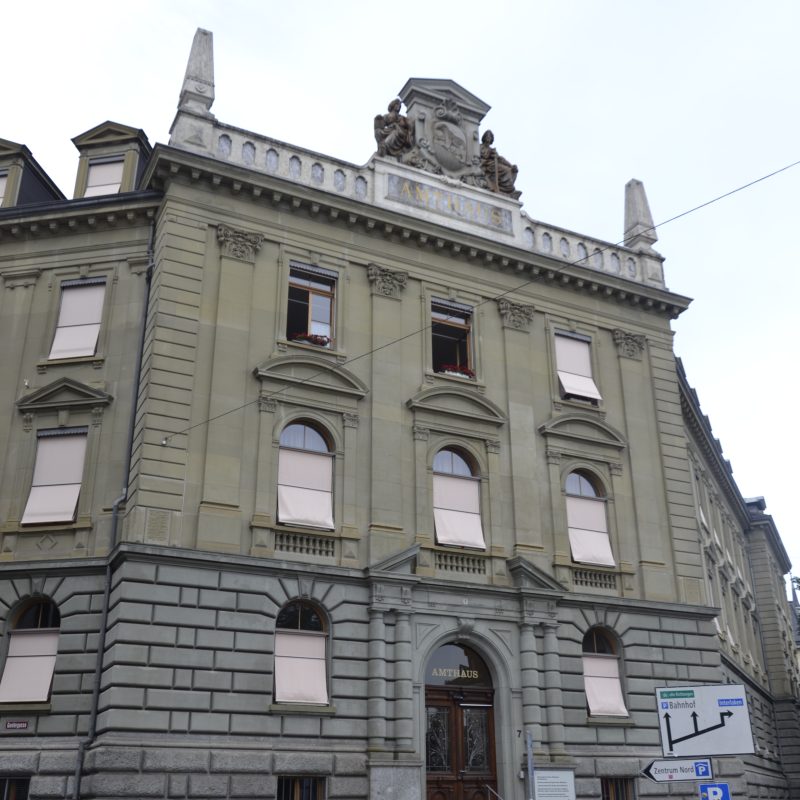 Le bâtiment de l'Amtshaus à Berne, conçu par l'architecte Franciszek Stempkowski, présente une façade néoclassique ornée de fenêtres cintrées et d'éléments décoratifs. L'entrée est marquée par un escalier menant à une porte en bois.