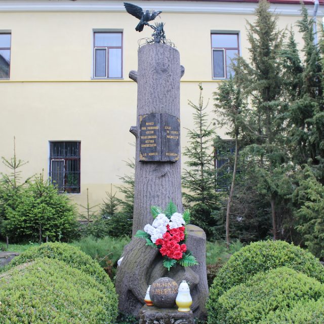 Pomnik Jerzego Wołodyjowskiego w Kamieńcu Podolskim. Pień drzewa z kapitelem i granitową kulą z napisem 'MAGNA RES LIBERTAS'. Na szczycie gołębica z gałązką. Tablica z inskrypcjami i kwiaty u podstawy.