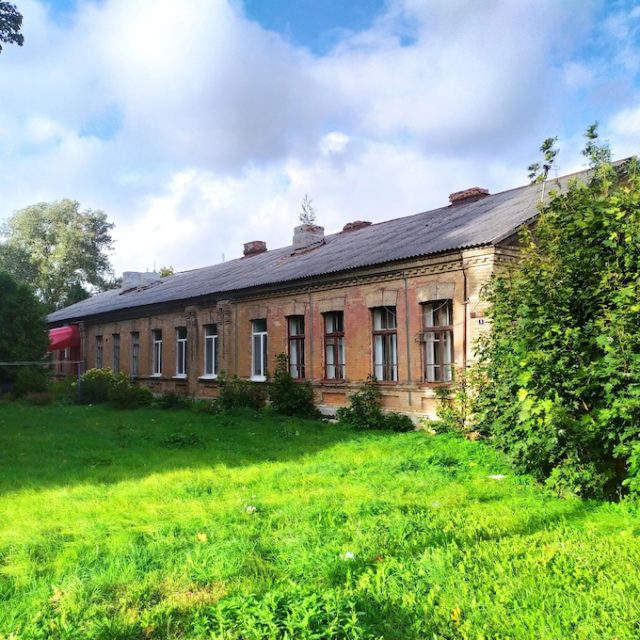 A historic building in Baranovichi, formerly the headquarters of the Novgorod Cavalry Brigade, with a long, single-storey structure, brick walls, surrounded by greenery.