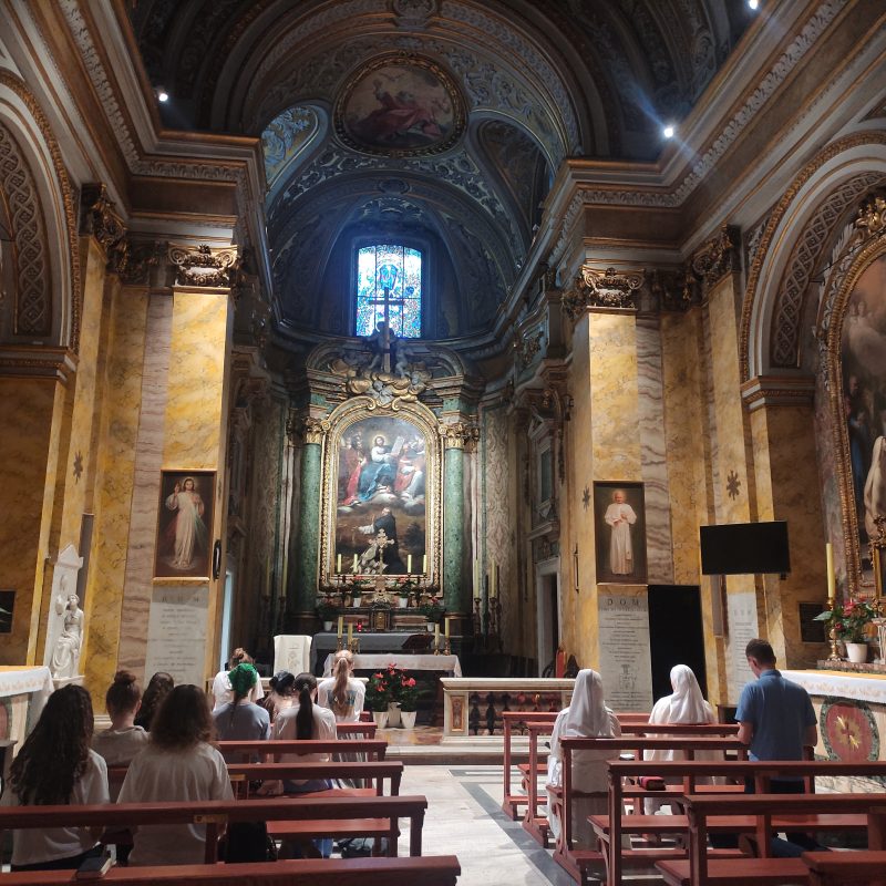 Intérieur de l'église Saint-Stanislas évêque et martyr à Rome, montrant l'autel avec des peintures religieuses et des personnes priant sur les bancs. L'architecture présente des arcs ornés et des éléments décoratifs.