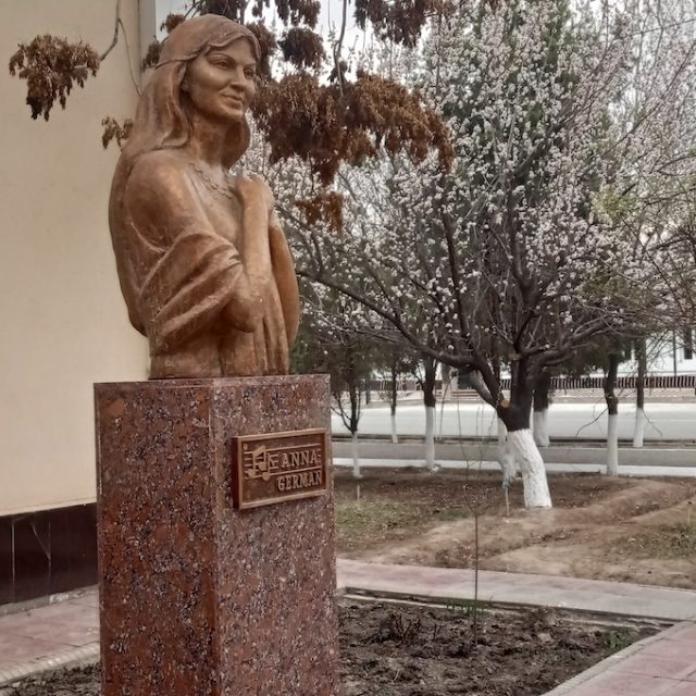 A bronze statue of Anna German on a granite plinth in Urgenchus, surrounded by trees. A plaque on the plinth bears her name.