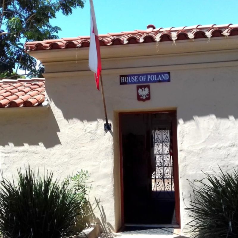 Dom Polski w Balboa Park w San Diego z polską flagą i tabliczką nad wejściem. Budynek ma dach z dachówek i jest otoczony zielenią.