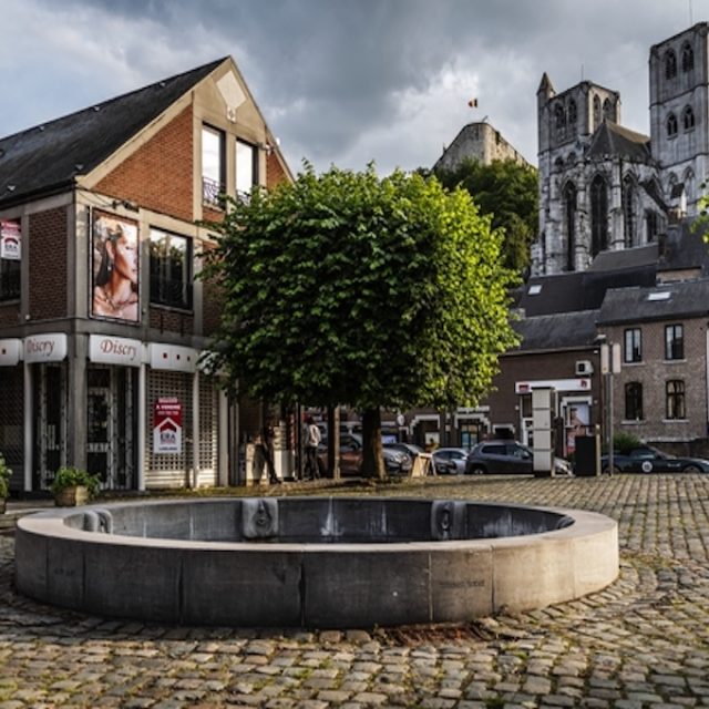 Granite fountain in the Saint-Severain square in Huy with a circular pool and jets shaped like throws. Surrounding cobblestone square with trees and buildings, including a church in the background.