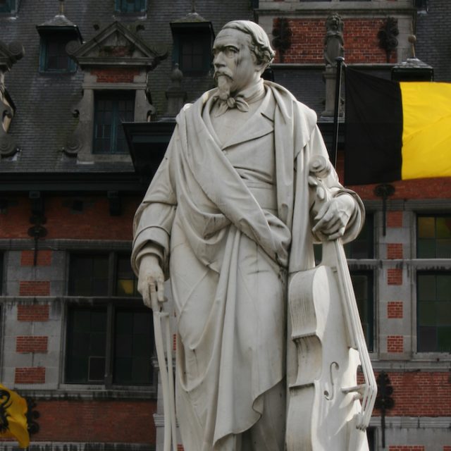 Statue of Adrien-François Servais holding a cello, on a pedestal with a laurel wreath, in front of a historic building with Belgian and Flemish flags in Halle.