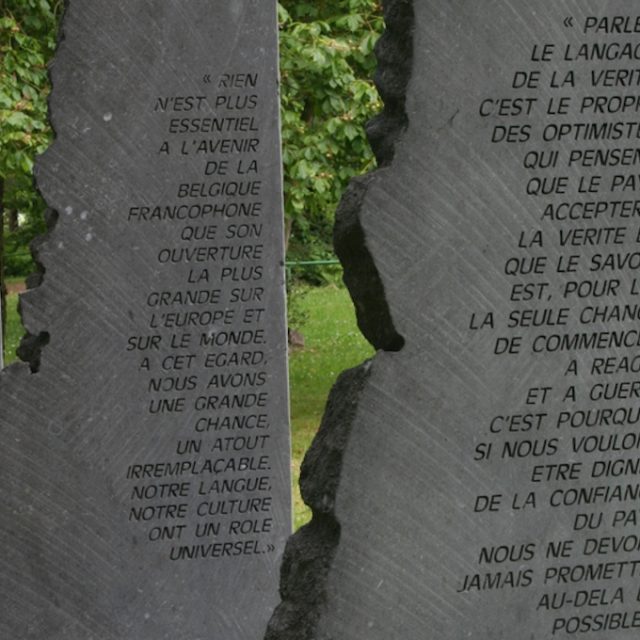 Four dark gray stone steles with engraved quotes from Jean Gol, located in a park in Embourg. The steles are thin, tall, and appear fragile, resembling wings.