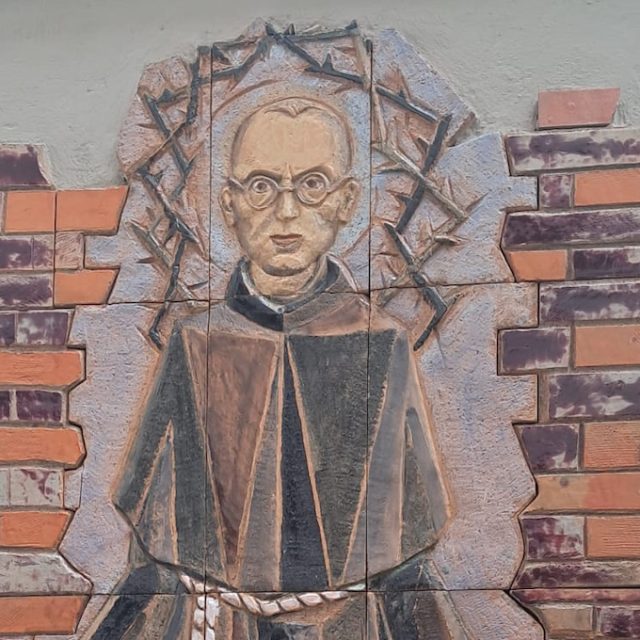 Terracotta relief of Maximilian Kolbe in a Franciscan habit with outstretched arms and a calm gaze, surrounded by stylised wall fragments and a crown of thorns, located in Mariahilferkirche, Graz.