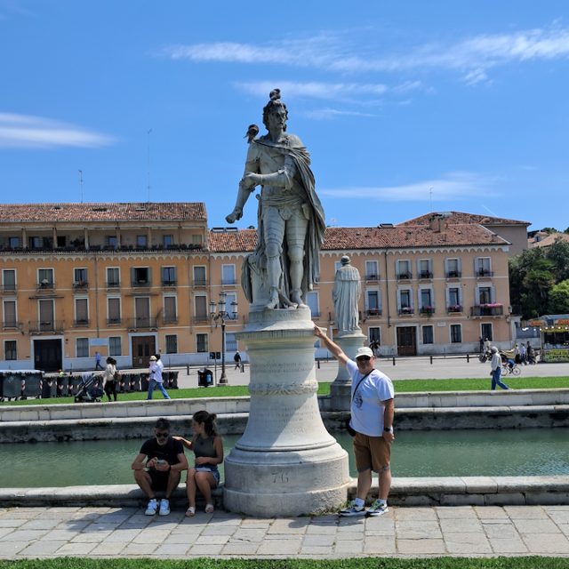 Pomnik Stefana Batorego na Prato della Valle w Padwie, przedstawiający postać w klasycznym stroju. W pobliżu siedzą i stoją ludzie. W tle zabytkowe budynki.