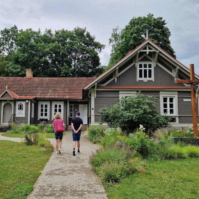 The wooden house at 29A Grybo Street in Vilnius, where St Faustina Kowalska lived. The building has a brown façade, a red roof and a cross in the garden. Two people walk along the path to the entrance.
