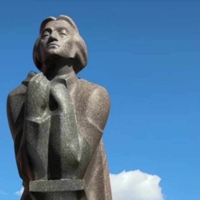 A granite monument to Adam Mickiewicz in Vilnius, near the Bernardine Church, with red-roofed buildings and a tree in the background.