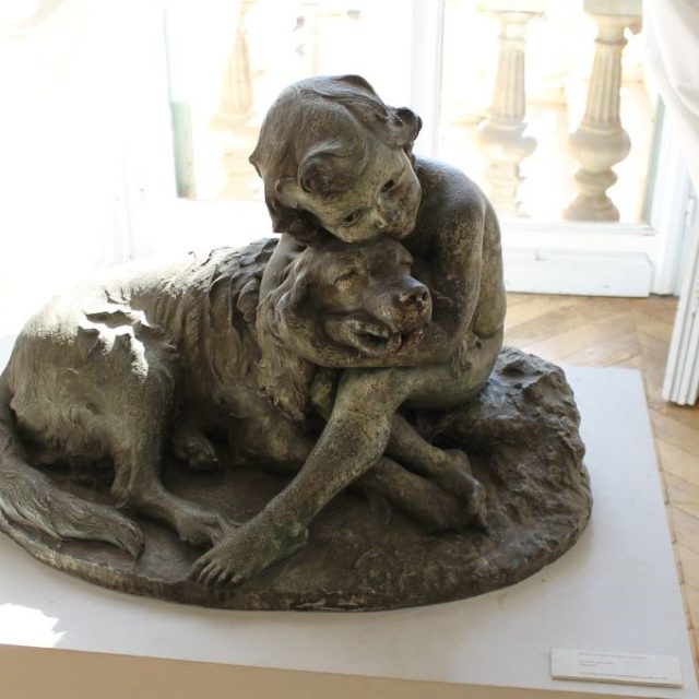 Brązowa rzeźba przedstawiająca dziecko obejmujące dużego psa, umieszczona na postumencie w pomieszczeniu z drewnianą podłogą i oknem w tle w Muzeum Sztuk Pięknych Julesa Chéreta w Nicei.