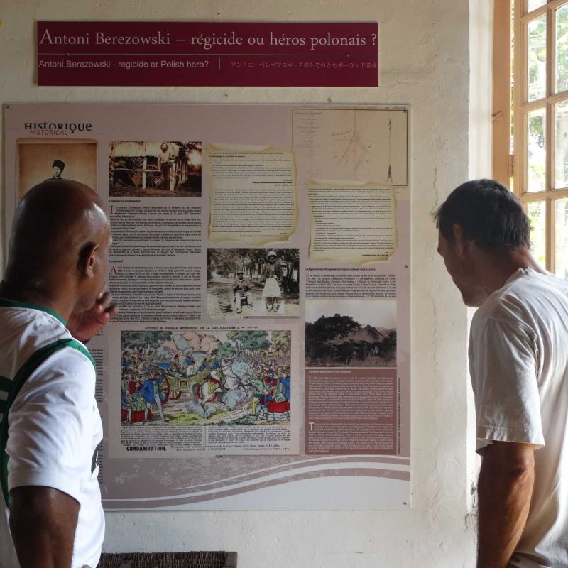 Two men look at an exhibition board about Antoni Berezowski at the Museum in Bourail, New Caledonia. The plaque contains text and historical photographs relating to Berezowski's life.