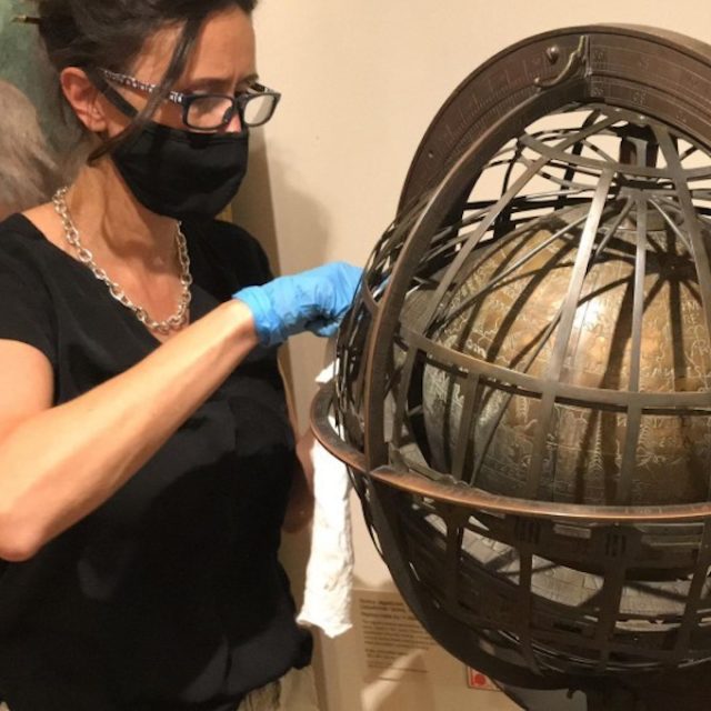 Konserwatorka w masce i rękawiczkach czyści dużą, ozdobną sferę armilarną w muzeum.