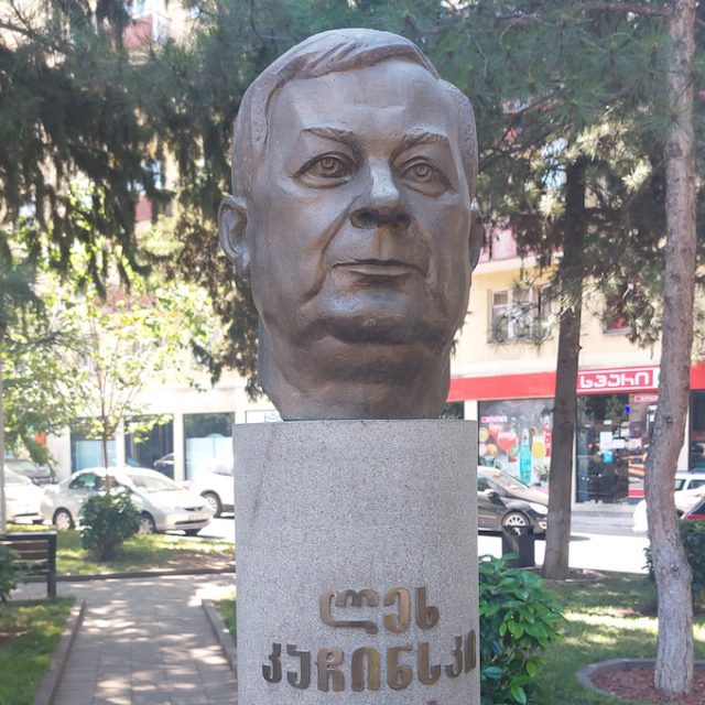 Brązowe popiersie Lecha Kaczyńskiego na cokole z gruzińskim napisem, otoczone drzewami i budynkami w Tbilisi.