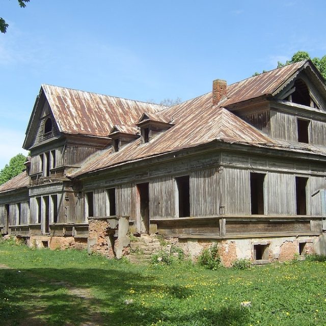 Ruins of the Rejtan Manor in Hruszówka, Belarus. The wooden building features a gabled roof with dormers and visible structural decay. The facade shows missing windows and deteriorating woodwork.
