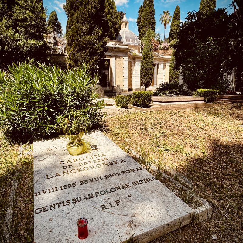 Pierre tombale de Carolina de Brezie Lanckoronska dans un cimetière, entourée d'arbres et de verdure. L'inscription comprend ses dates de naissance et de décès, et une bougie rouge est placée sur la tombe.