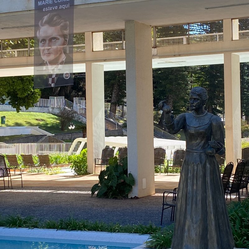 La statue de Marie Skłodowska-Curie à Águas de Lindóia, debout près de la piscine sous une structure moderne. Une bannière avec son portrait et l'inscription "Marie Curie esteve aqui" est suspendue au-dessus de la statue.
