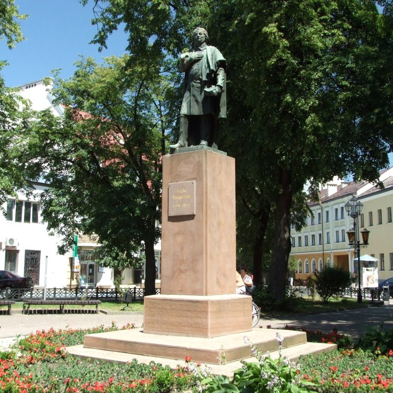 Pomnik Adama Mickiewicza na cokole w otoczeniu drzew i budynków w parku w Stanisławowie.