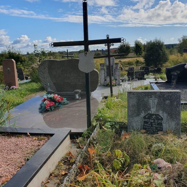 Graves of World War II victims in Góże village cemetery. A cross and tombstones with inscriptions are visible, surrounded by grass and flowers under a cloudy sky.