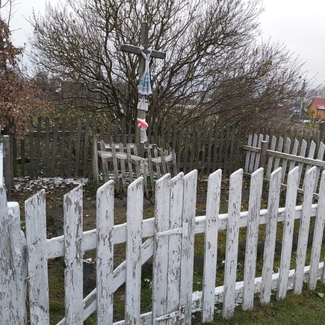 Tombe d'un légionnaire tombé en 1915, entourée d'une clôture en bois blanc. Une croix avec un drapeau polonais et une écharpe est visible. Arbres nus et paysage rural en arrière-plan.