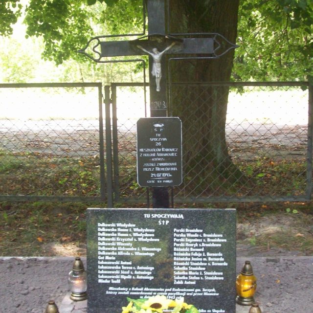 Grave of 26 inhabitants of Rudka murdered by UPA in 1943, marked by a cross with a crucifix and a plaque with names. Surrounded by a fence and trees, with candles and sunflowers at the base.