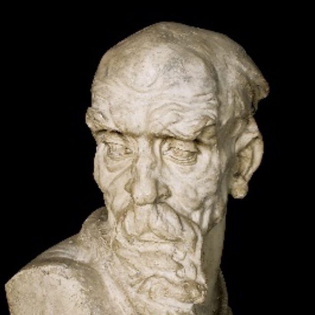 Sculpture représentant la tête d'un homme âgé aux traits marqués, notamment des yeux enfoncés et une moustache bien dessinée, sur fond noir.