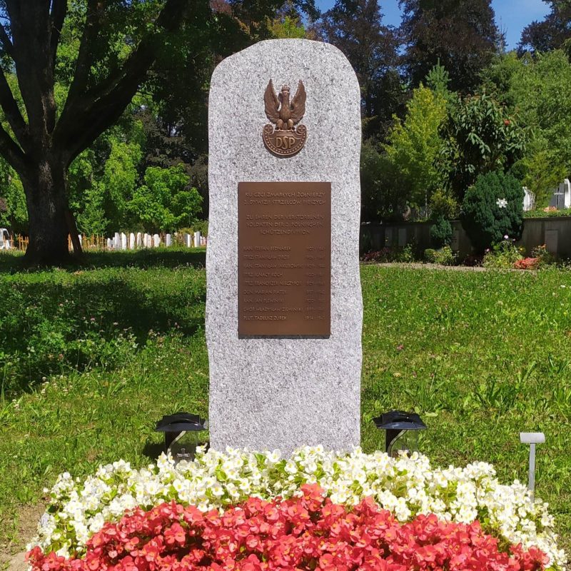Granitowy obelisk z tablicą upamiętniającą żołnierzy 2. Dywizji Strzelców, otoczony kwiatami i zielenią na cmentarzu.