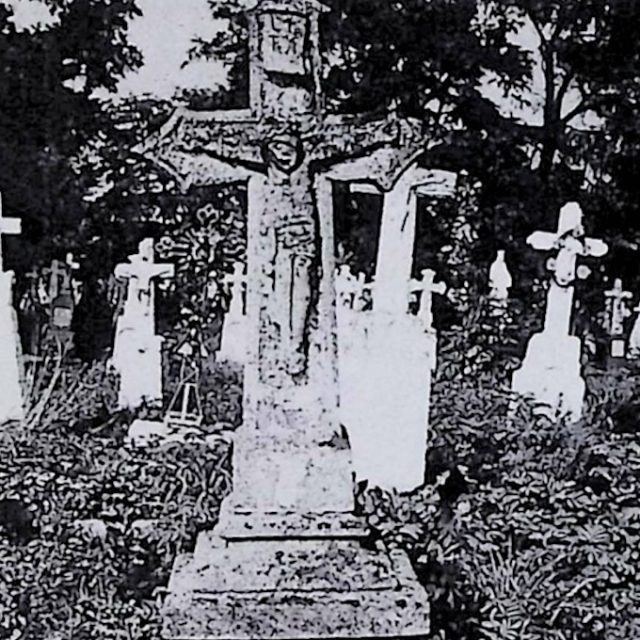 Gravestone with a cross on a two-tiered pedestal. The cross features a relief of Christ and an inscription. Surrounding are other gravestones amidst vegetation.