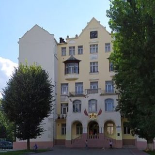 Fotografia wielokondygnacyjnego budynku we Lwowie z centralnym wejściem i arkadowymi oknami, wykuszem nad wejściem oraz flagami nad drzwiami, w otoczeniu drzew