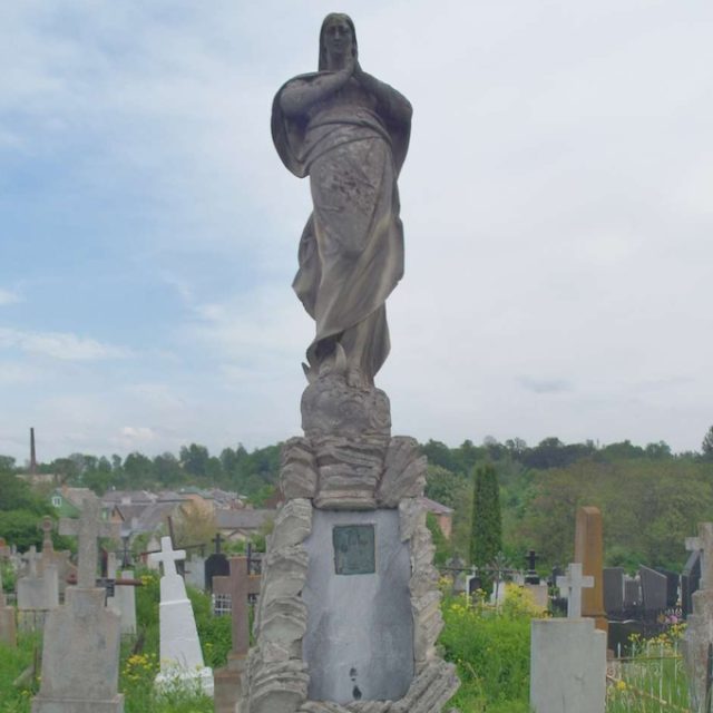 Photographie d'une pierre tombale ornée d'une statue de la Vierge Marie posée sur un socle imitant des rochers et reposant sur une base à trois marches comportant une plaque commémorative