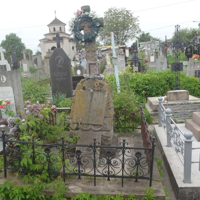 Photographie d'une tombe dans un cimetière entourée d'une clôture en fonte ornée de niches en forme de lys, avec une croix en pierre en forme de tronc recouvert de lierre, un socle orné d'une ancre et d'un livre ouvert comportant une inscription, ainsi qu'une plaque inclinée gravée