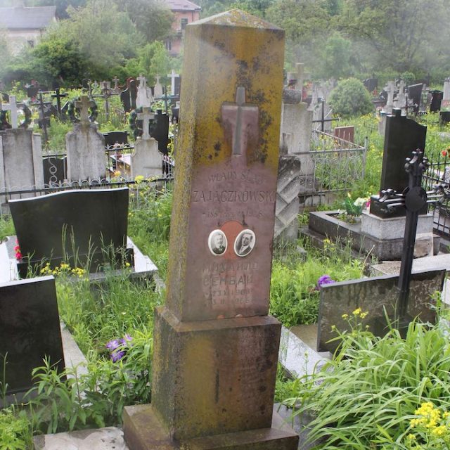 Photographie d'une tombe dans un cimetière, comportant un obélisque en pierre posé sur un socle, orné d'une croix gravée et d'une inscription, ainsi que de deux portraits ovales en porcelaine sur la façade