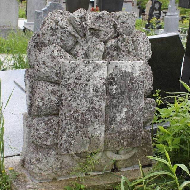 Pierre tombale dans le cimetière en forme de rochers avec un bas-relief représentant un livre ouvert encadré d'une ancre et de fleurs au-dessus et une inscription gravée sur les pages.