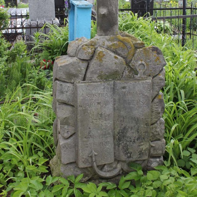 Photographie d'une pierre tombale en pierre avec une croix stylisée en tronc d'arbre, sur un socle de rochers et un livre ouvert avec une ancre, avec des motifs végétaux gravés et une inscription sur les pages.