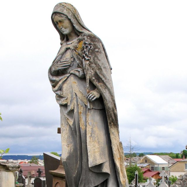 Photographie d'une statue de cimetière représentant la Vierge Marie sur un piédestal en forme de rocher, la tête inclinée, la main droite sur la poitrine, la main gauche tenant un lys, et une plaque d'inscription sur le devant.