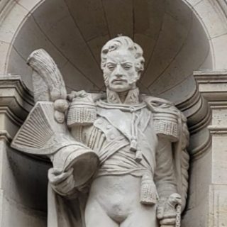 A stone sculpture of Prince Józef Poniatowski in a niche on the façade of the Louvre in Paris, wearing a uniform with a cape, holding a shako with a tassel and resting his hand on the hilt of his sabre