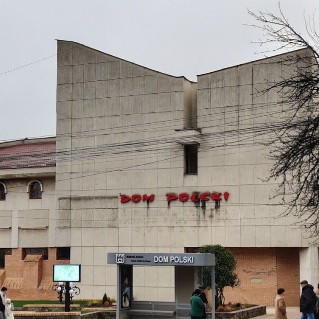Photo du bâtiment « Dom Polski » à Suczawa, avec l'inscription « Dom Polski » en rouge sur la façade et des passants dans la rue.
