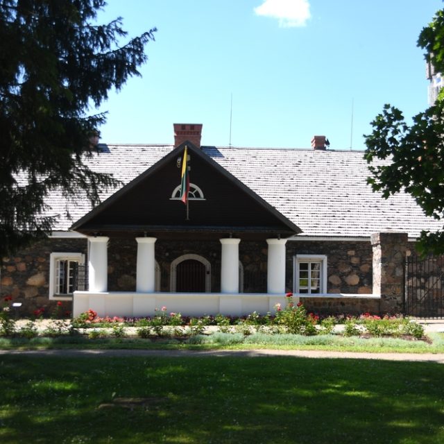 Stone manor house in Burbiskis with a columned porch, dark roof, flower beds and trees in the park.