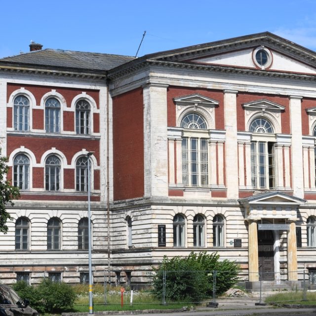 Le bâtiment de l'ancien gymnase Mikołajowski à Lipawa, avec sa façade en briques rouges, ses pilastres blancs, ses hautes fenêtres en demi-cercle et son portique d'entrée à colonnes.