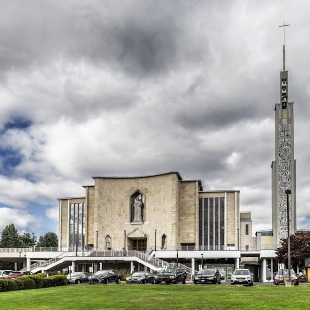 Fotografia Narodowego Sanktuarium Matki Boskiej Częstochowskiej w Doylestown z szeroką fasadą kościoła, schodami, zaparkowanymi samochodami i wysoką dzwonnicą zwieńczoną krzyżem.