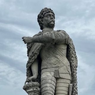 Une statue en pierre à Padoue représente un homme en armure et en manteau, la main posée sur une couronne, sur fond de ciel nuageux.
