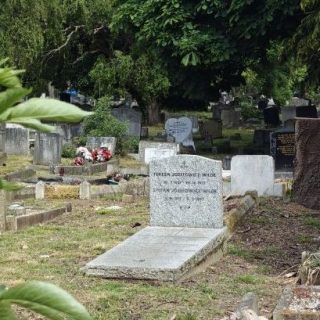 Granitowy grób z poziomą płytą i pionową stelą na cmentarzu South Ealing w Londynie pośród innych nagrobków i drzew.