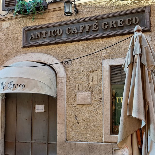 Fotografia wejścia do Antico Caffè Greco w Rzymie z szyldem nad drzwiami i napisem Caffè Greco na markizie.